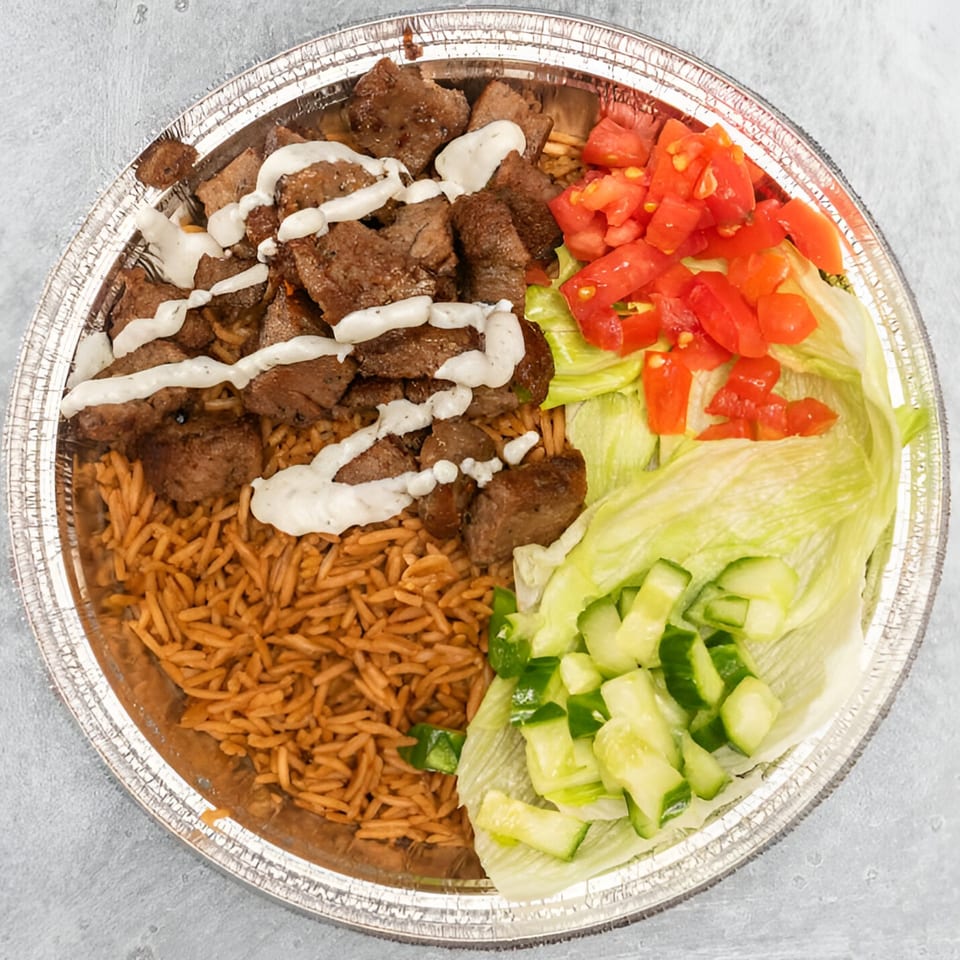 Halal beef and rice platter with white sauce, diced tomatoes, lettuce, and cucumbers in a foil container.