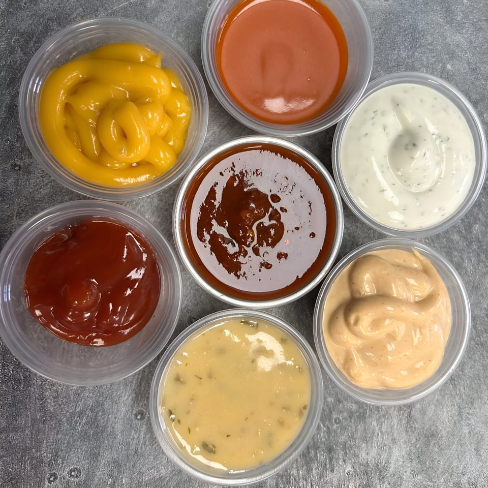 Seven small plastic cups of various sauces, including mustard, ketchup, BBQ, and creamy dressings.