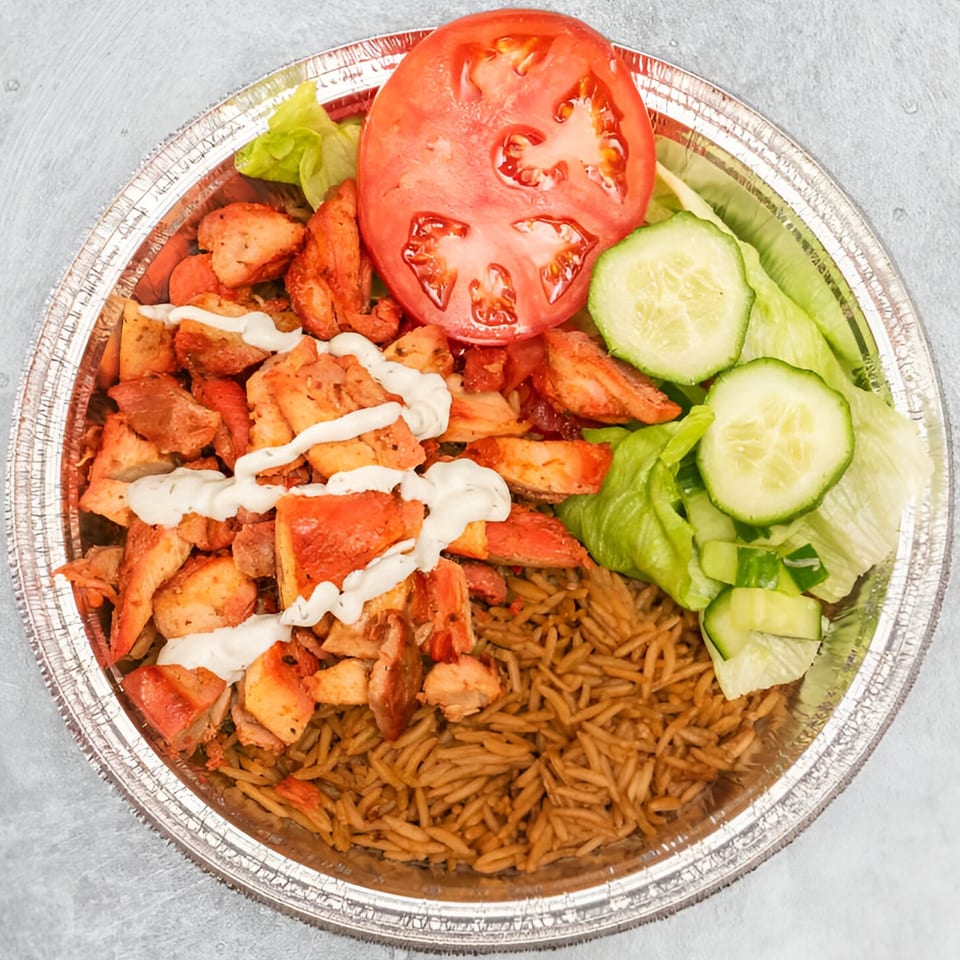 Halal chicken over rice with white sauce, sliced tomatoes, and cucumbers in a foil container.