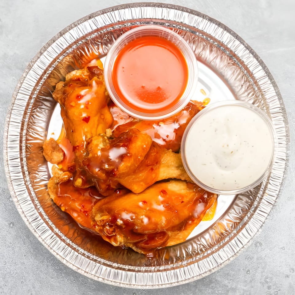 Sweet chili chicken wings with a side of ranch and hot sauce in a foil container.